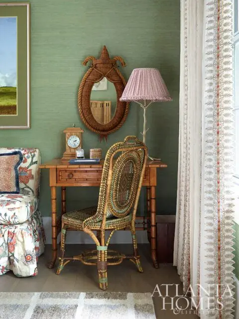 Green grasscloth wallpaper with a wooden desk, wicker chair, and patterned armchair.