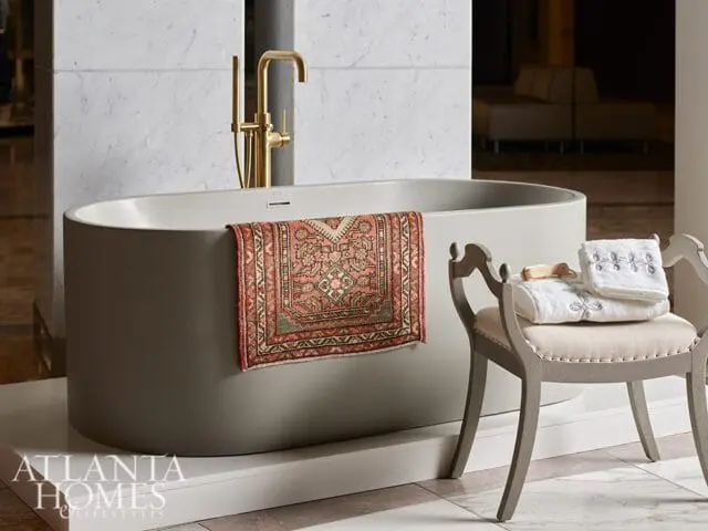 Gray freestanding bathtub with gold faucet, small Persian rug, and vanity stool with towels