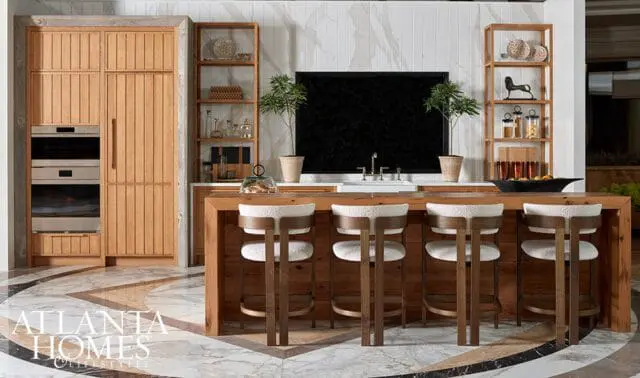 Wood kitchen with paneled refrigerator, island with white upholstered stools, and marble floor.
