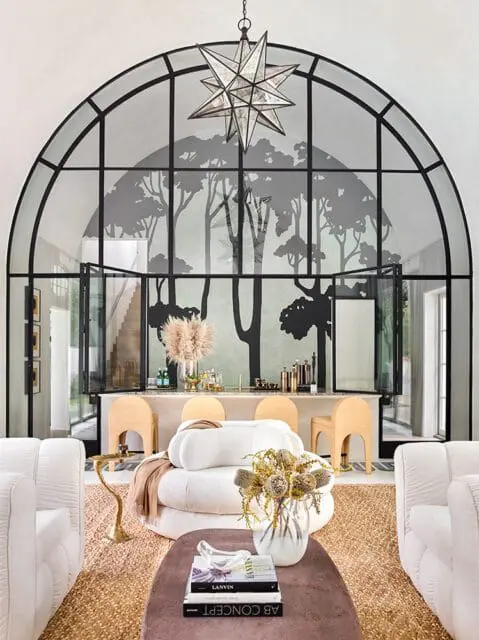 Living room with a large arched window featuring a black tree silhouette mural, a bar area with rattan stools, and white tufted sofas.