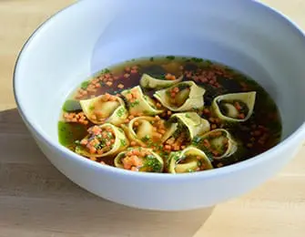 White bowl filled with broth, tortellini pasta, and finely diced carrots and herbs.