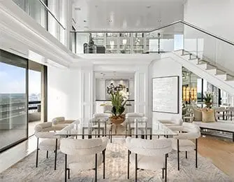 Modern dining room with glass table, plush white chairs, and a floating staircase with glass railings. White walls and built-in shelving create an airy feel.