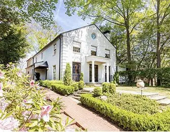 White stucco house with dark trim, arched entryway, and circular window above. Landscaped garden with manicured hedges and brick pathway.