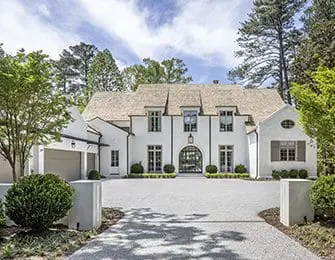 White stucco French country home with arched entryway, dormer windows, and attached garage, set on a gravel driveway with manicured landscaping.