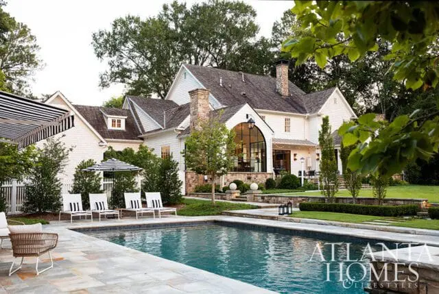 White farmhouse-style home with stone accents, large arched window, and a swimming pool in the foreground.