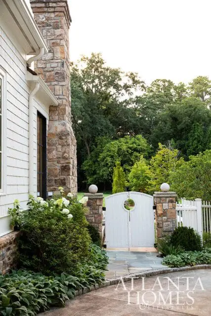 White arched gate with circular cutout, flanked by stone pillars topped with spheres, leading to a lush garden with mature trees.