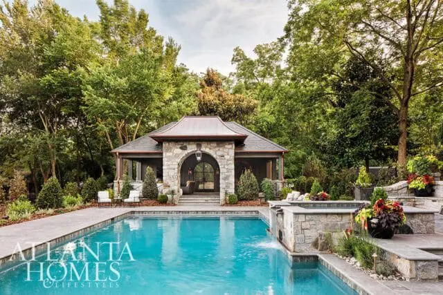 Luxury backyard with a bright blue swimming pool and spa, stone-clad pool house with arched entryway, and lush green trees.