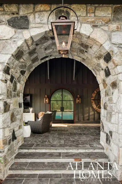 Stone archway entrance with a hanging lantern, leading to a covered patio with dark wood paneling, a large arched window, and a glimpse of a swimming pool.