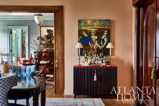 Dining room with a dark wood table and blue tufted sofa, opening to a living area with a bar cabinet displaying bottles and a painting of three figures.