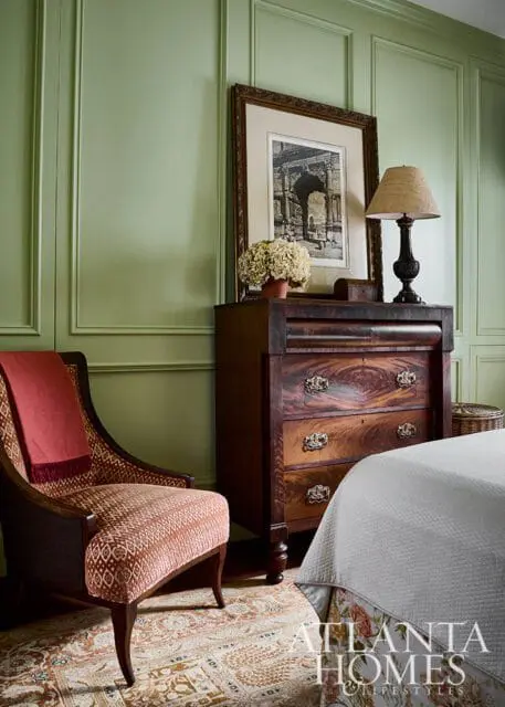 Bedroom with light green paneled walls, a patterned armchair with a coral throw, an antique mahogany dresser, and a framed print above a table lamp.