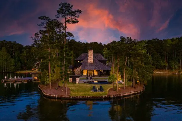 Lakefront home with stone chimney and dark wood siding, surrounded by pine trees and a dock, under a dramatic twilight sky with orange and purple clouds.