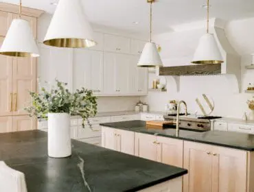 White kitchen with light wood cabinetry, dark stone countertops, brass hardware, and large pendant lights with brass interiors.