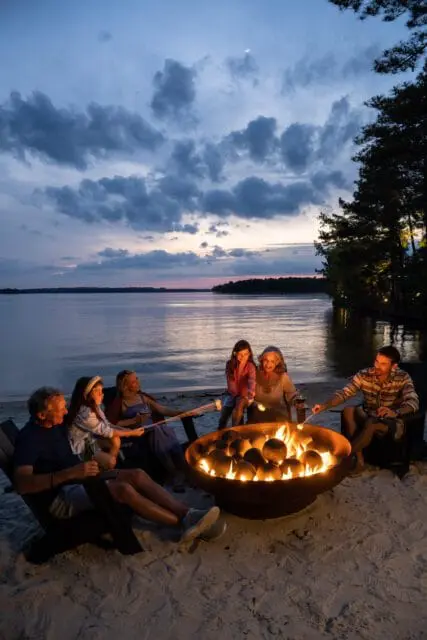 Family roasting marshmallows over a fire pit on a sandy beach at dusk, with a lake and trees in the background.