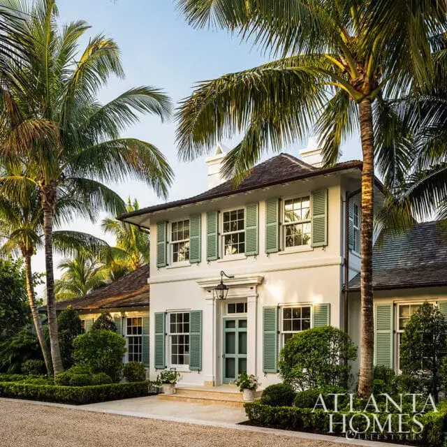White two-story home with light green shutters, a dark roof, and a light blue door, surrounded by lush landscaping and palm trees.