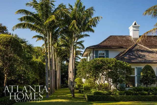 White house with light blue shutters and a wooden shake roof, surrounded by lush greenery and tall palm trees.
