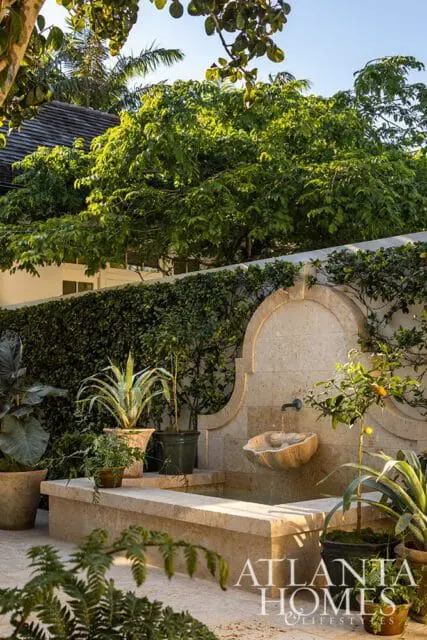 Stone fountain with a scalloped basin and arched surround, surrounded by lush greenery and potted plants.