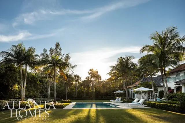 Luxury backyard with a swimming pool, lounge chairs, umbrellas, and palm trees under a bright blue sky with sunbeams.