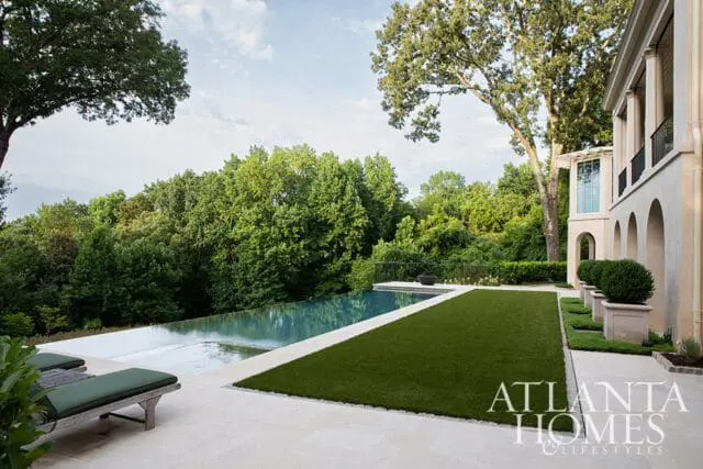Infinity pool with a stone patio and manicured lawn, surrounded by lush trees. The side of a stucco home with arched windows is visible.