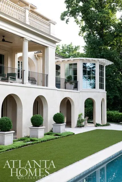Cream stucco house with arched loggias, balconies, and a sunroom addition. Landscaped grounds feature manicured hedges, topiary balls in planters, and a swimming pool.