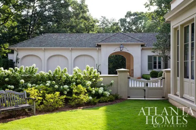 Cream-colored garage with arched doors and a covered entryway, flanked by lush green landscaping and a white picket fence and gate.