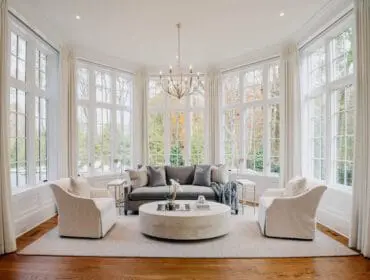 Living room with a gray sofa, two white armchairs, a large round concrete coffee table, and floor-to-ceiling windows overlooking autumn trees.