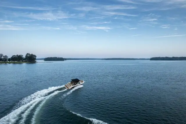 Motorboat speeding across a wide blue lake, leaving a white wake, with tree-lined shores and islands in the distance under a pale blue sky with wispy clouds.