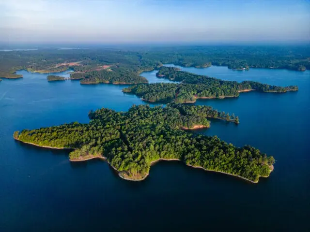 Aerial view of a large, tree-covered island in a deep blue lake, with smaller islands and a distant shoreline visible under a clear sky.