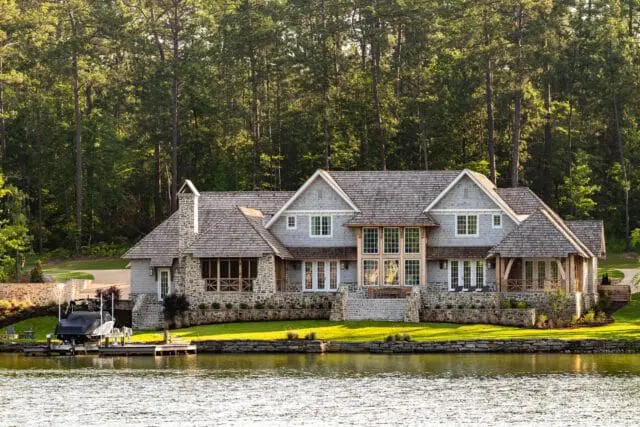 Large stone and shingle lake house with multiple gables, large windows, and a covered boat dock in the foreground.