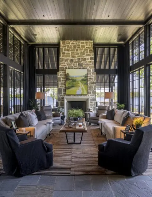 Living room with stone fireplace, large windows overlooking a lake, dark wood ceiling, and seating arranged around a striped rug and wooden coffee table.