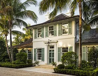 Two-story white house with light green shutters and a dark shingled roof, surrounded by lush landscaping and palm trees.