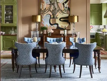 Dining room with a dark wood table and chairs upholstered in blue and white patterned fabric, flanked by green built-in cabinetry. A colorful abstract painting hangs above a buffet.