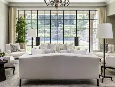 Living room with cream upholstered sofas and armchairs, patterned throw pillows, a large black-paned window overlooking a garden, and a traditional chandelier.