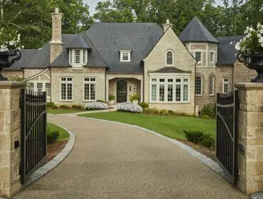 Stone estate with dark grey roof, arched windows, and a winding driveway leading to a black metal gate flanked by stone pillars with lanterns and urns