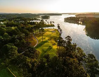 Aerial view of a golf course on a peninsula jutting into a large lake at sunrise, with pine trees and rolling green fairways.