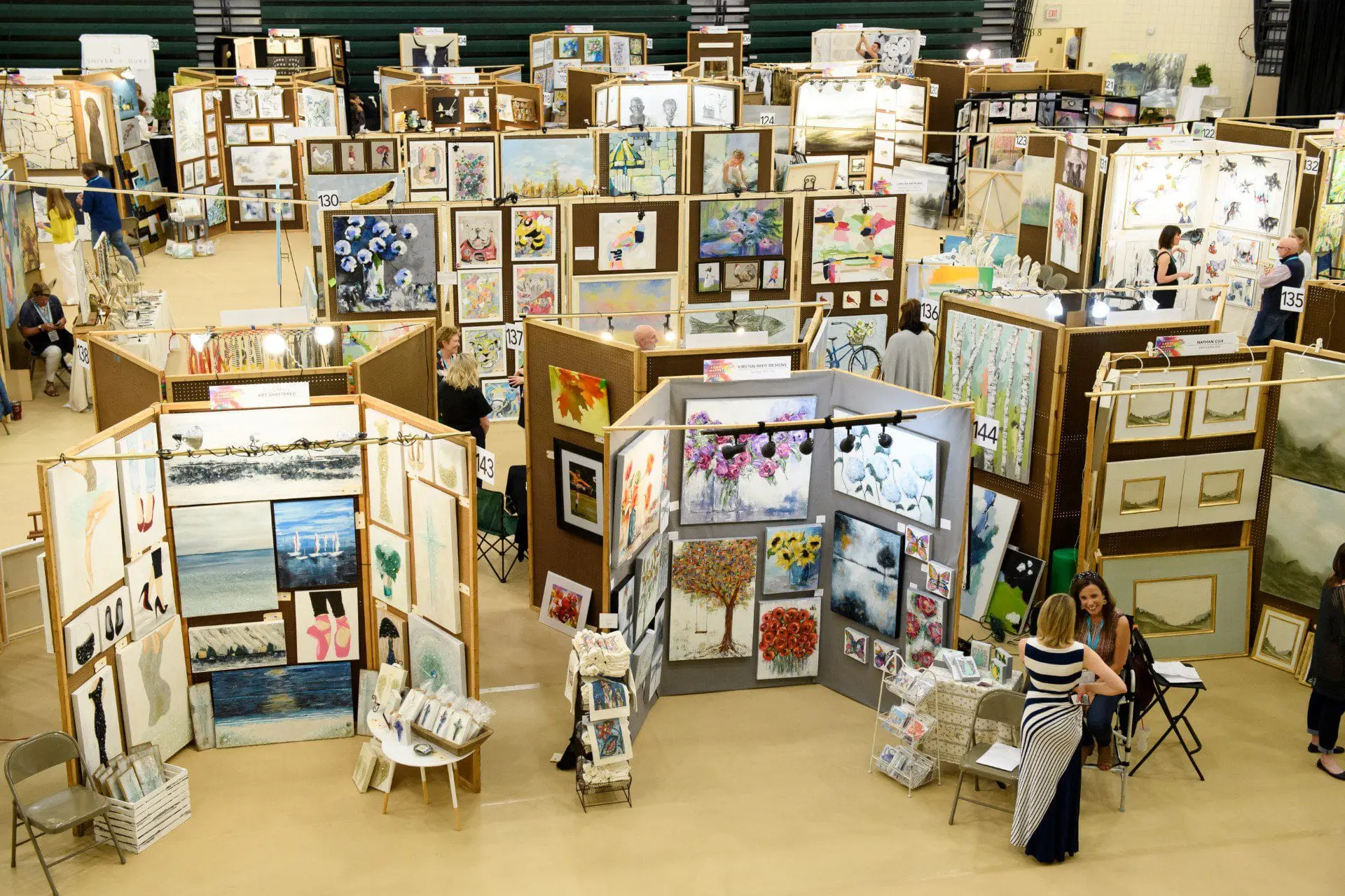 Art exhibition with numerous booths displaying paintings and artwork, featuring various styles including landscapes, portraits, and abstract pieces. People are browsing the displays.