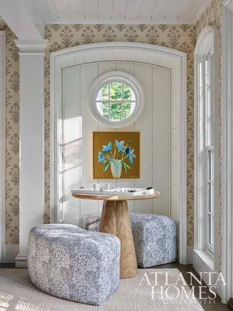 Entryway with patterned wallpaper, a round window, a wooden pedestal table with ottomans, and a painting of blue flowers.