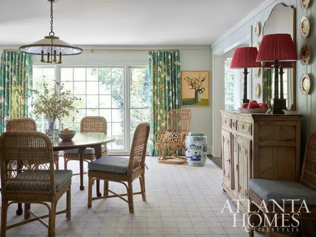 Dining room with wicker chairs around a wooden table, botanical print curtains, and a vintage wooden cabinet with red lamps.