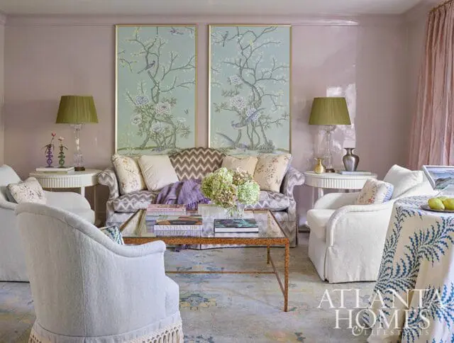 Living room with pale pink walls, featuring a patterned sofa, two upholstered armchairs, a glass-top coffee table, and two framed botanical prints with birds and flowers.