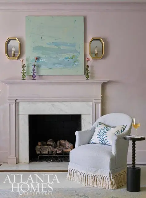 Light blue armchair with a blue and white patterned pillow in front of a fireplace with a marble surround. Above the fireplace is a large abstract painting and two brass sconces.