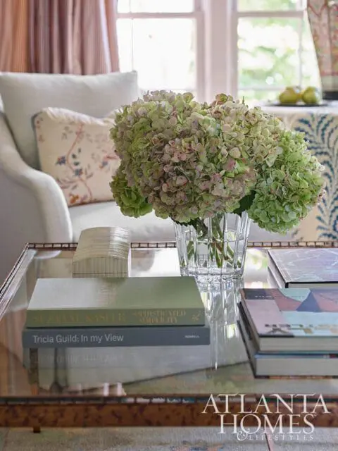 Living room interior with a glass coffee table holding books and a vase of hydrangeas, with a chair and window in the background.