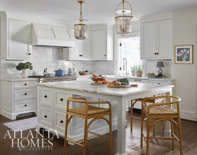 White kitchen with marble countertops and backsplash, custom white cabinetry, a large island with seating, and brass pendant lights.