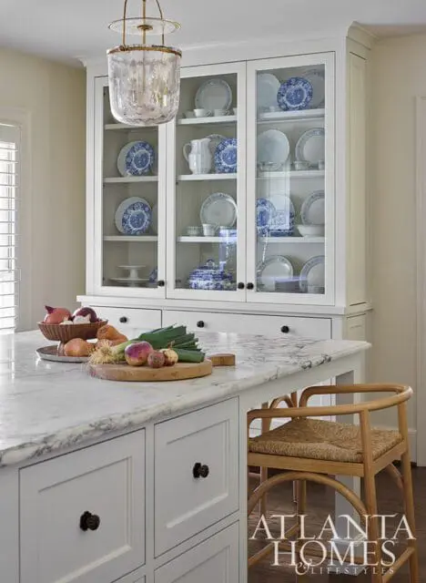 White kitchen island with marble countertop, white cabinetry, and a glass-front hutch filled with blue and white dishes. A wooden stool is tucked under the island.
