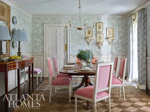 Dining room with light blue floral wallpaper, a dark wood table set for six, pink upholstered chairs with white frames, and a mirrored console table with lamps.