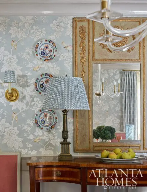 Light blue floral wallpaper with birds, a brass sconce, decorative plates, a table lamp with a patterned shade, and a wooden console table with a tray of pears.