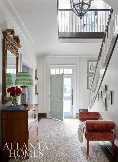 Entryway with a wooden chest, ornate mirror, green lamp, red flowers, light blue door, and red upholstered bench. A staircase with a lantern hangs overhead.