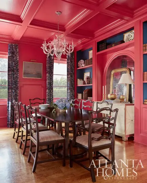Dining room with bright coral walls and coffered ceiling, dark wood dining table and chairs, and navy blue built-in bookshelves.
