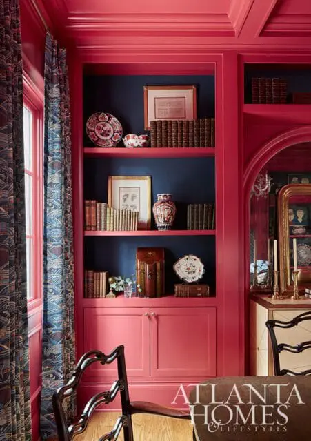 Dining room with fuchsia built-in bookshelves and cabinets, dark blue walls, patterned curtains, and antique chairs.