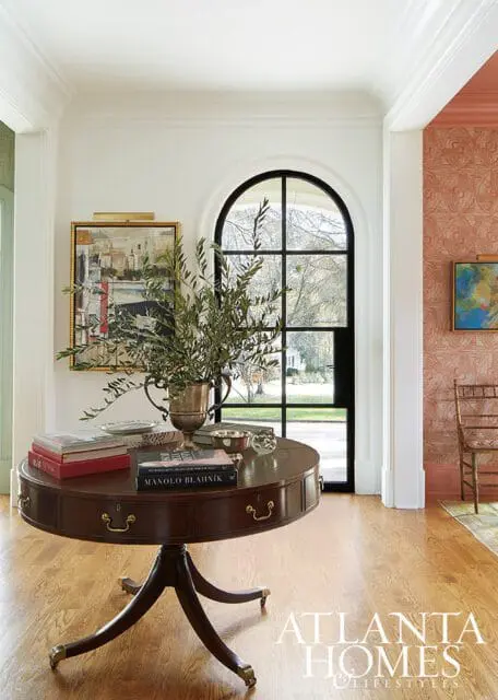 Foyer with a round mahogany pedestal table topped with books and a silver vase of greenery, beneath an arched window overlooking a lawn.