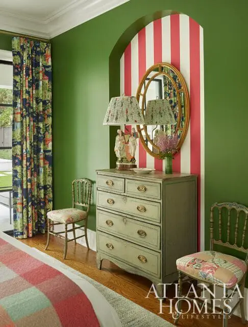 Green walls in a bedroom with a sage green dresser, a gold oval mirror, and two chairs upholstered in floral fabric.