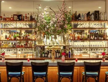 Bar with wooden cabinetry, marble countertop, and shelves stocked with liquor bottles and glassware. Four blue upholstered bar stools face the bar.